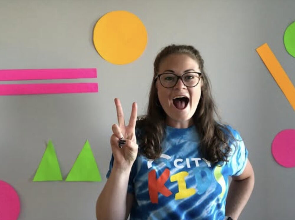 Leah in a City Kids tie-dye shirt, flashing a peace sign in front of a wall of colourful paper shapes